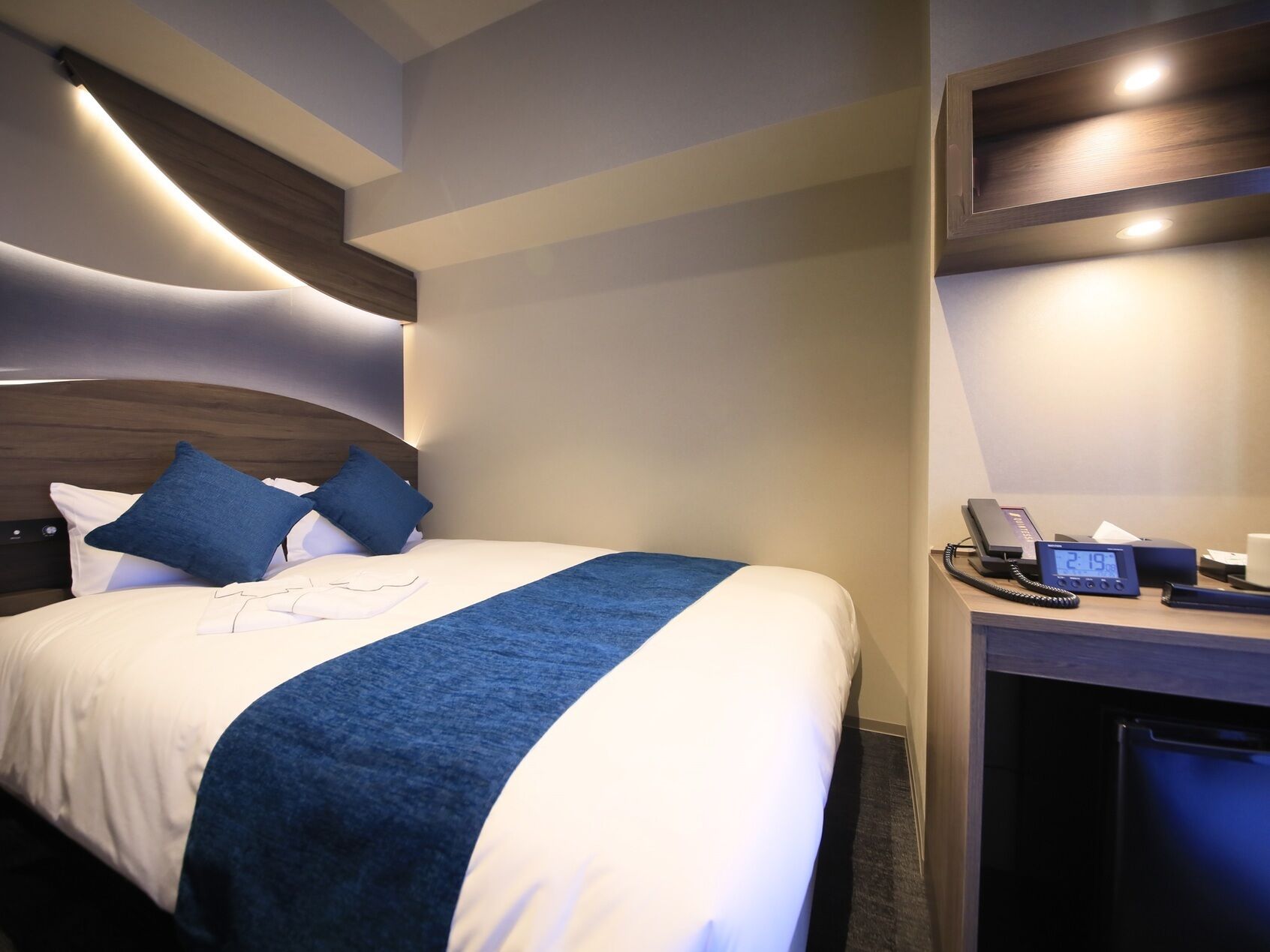 Une chambre d’hôtel comprenant un lit, un bureau avec un téléphone et une horloge numérique, ainsi qu’un mini-bar.