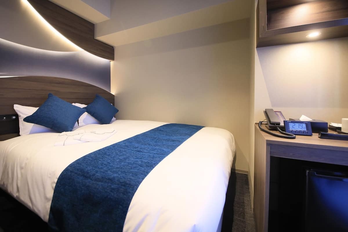 Une chambre d’hôtel comprenant un lit, un bureau avec un téléphone et une horloge numérique, ainsi qu’un mini-bar.