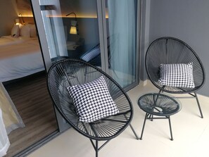 Twee zwarte stoelen met draadframe en hondentand patroon kussens, een klein rond tafeltje, en een bed met witte beddengoed in een moderne kamer met een glazen deur en uitzicht.