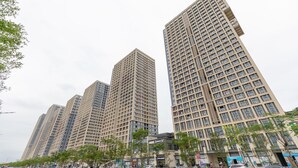 Hohe Wohngebäude mit einer rasterförmigen Fassade, umgeben von Bäumen und einigen Geschäften.