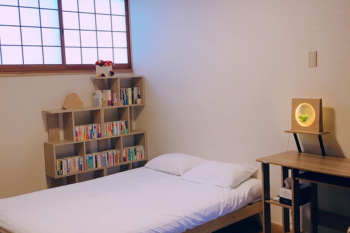A small, neatly arranged bedroom with a single bed, a wooden nightstand, a bookshelf, and a window with blinds.