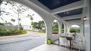 A covered outdoor area with white chairs and tables, a tiled floor, and a view of greenery and a building in the background.