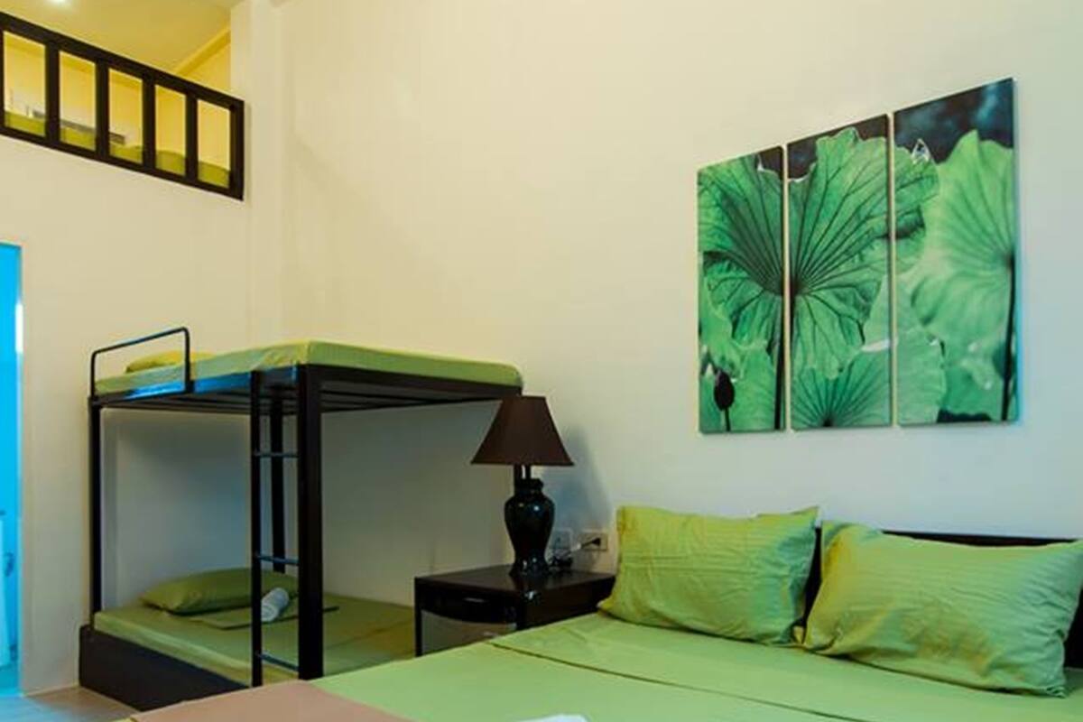 Une chambre d’hôtel avec un lit superposé, un lit simple, une couvre-lit verte, une couverture marron, une lampe et une décoration murale représentant des feuilles vertes.