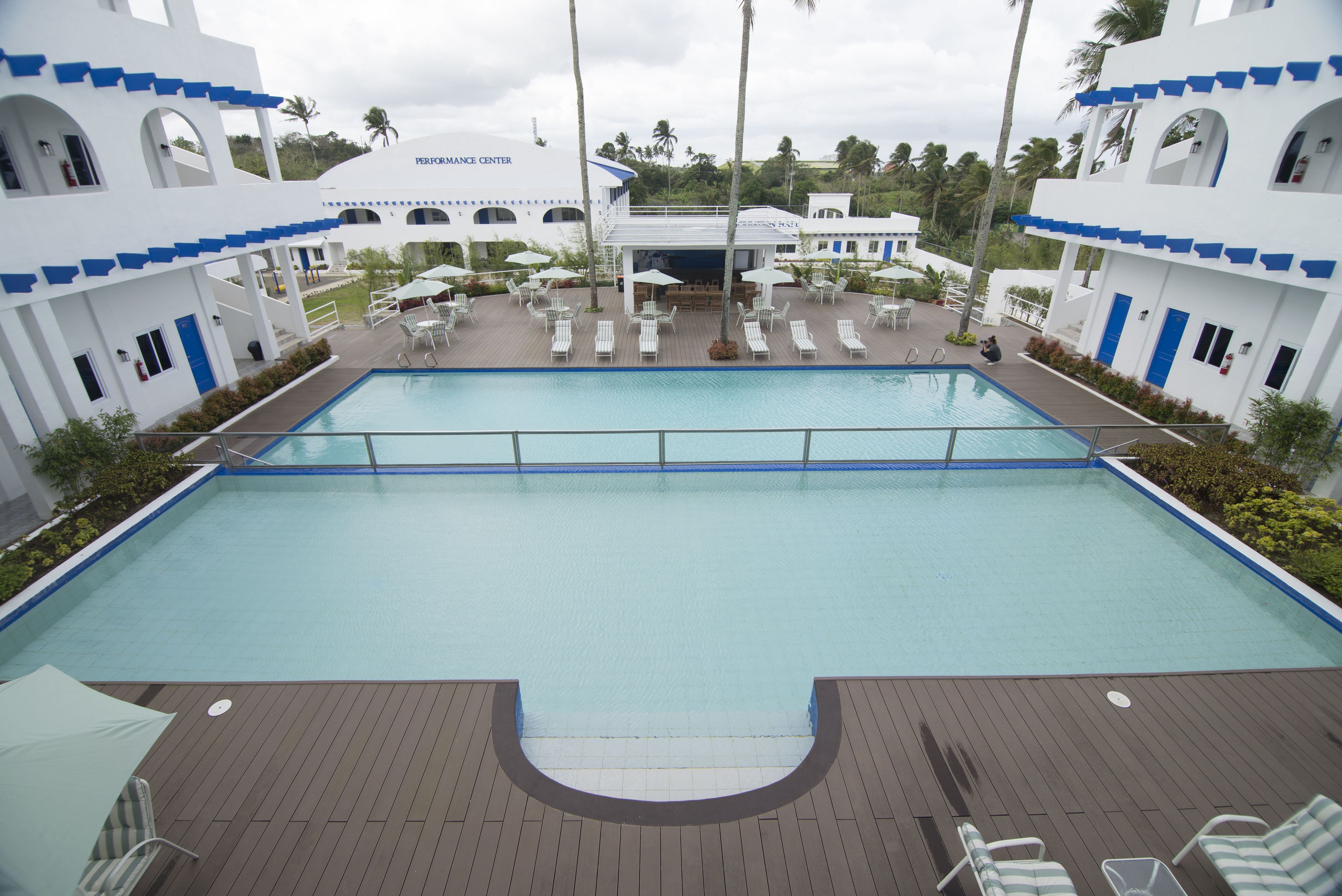 A large swimming pool with lounge chairs and umbrellas, surrounded by white buildings with blue accents and palm trees.