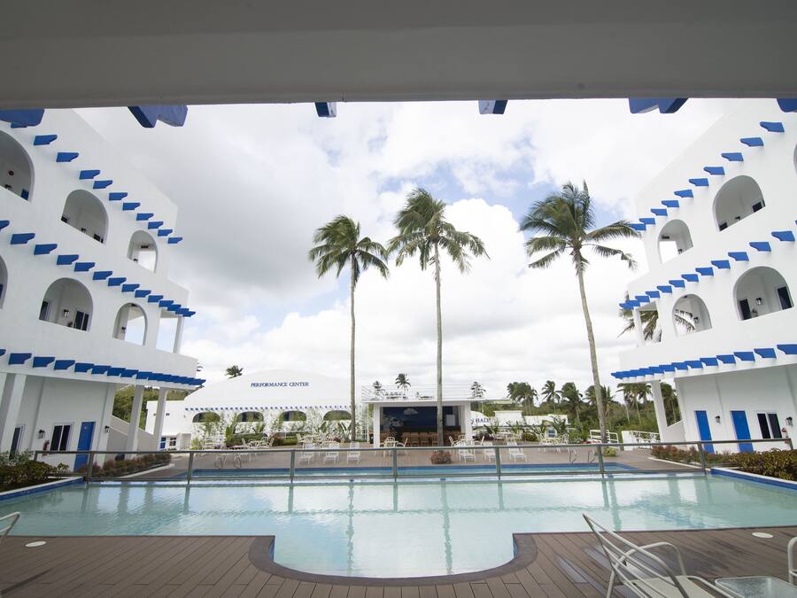 A swimming pool with a wooden deck, surrounded by white buildings with blue accents and palm trees.