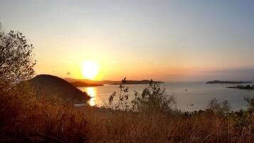 A coastal landscape with a sunset over water, hills, and sparse vegetation.
