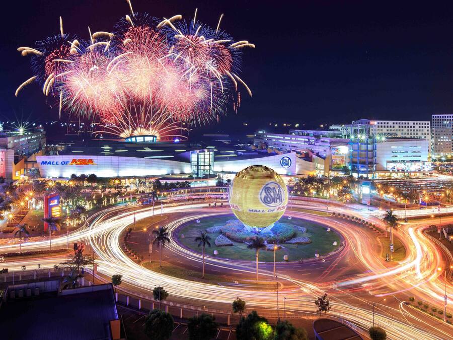 Paesaggio urbano notturno con una piazza circolare ben visibile, un grande globo illuminato e un edificio con la scritta "MALL OF ASIA".