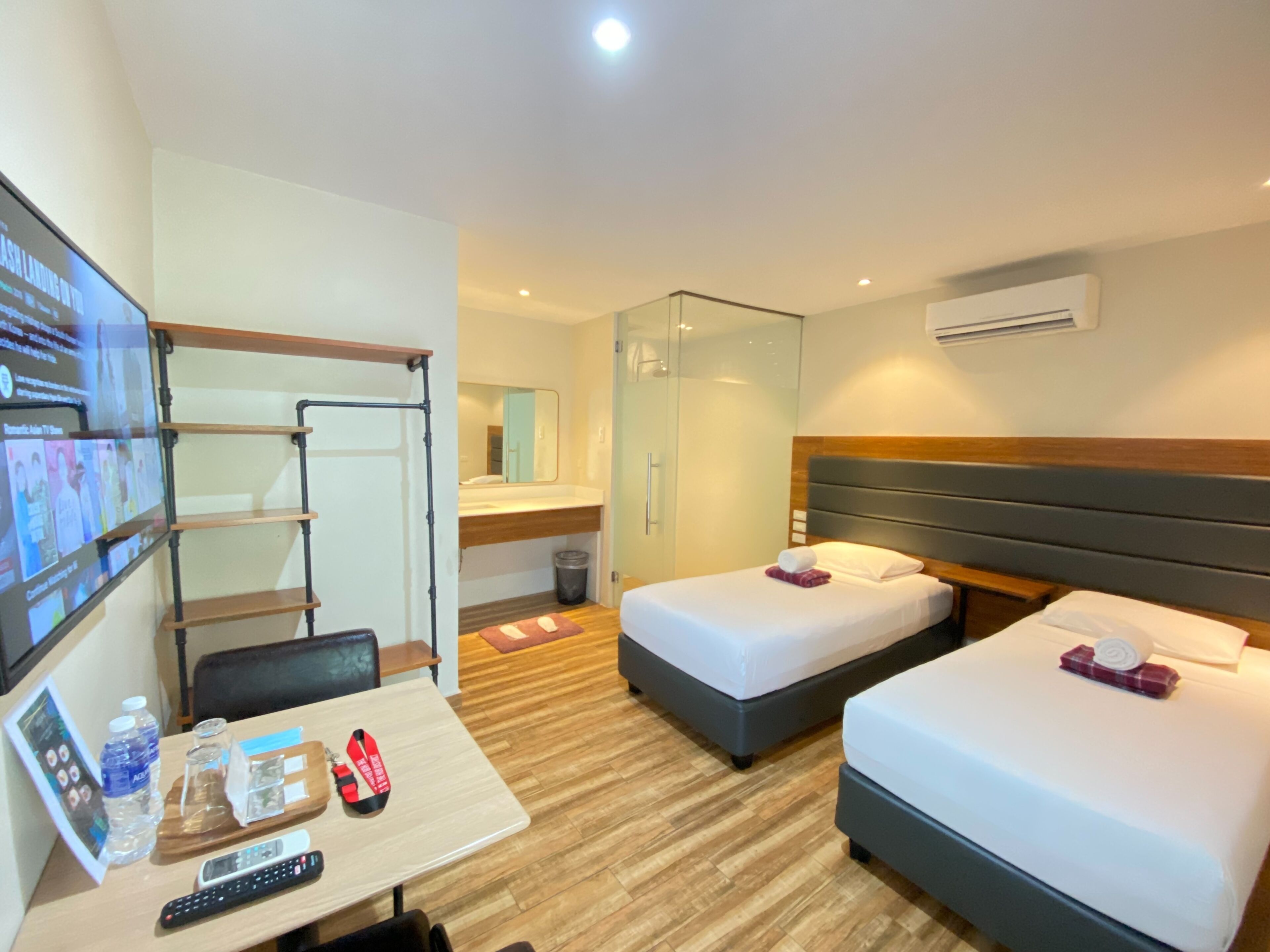 Quarto de hotel com duas camas, uma escrivaninha com televisão, um espelho e área de chuveiro.