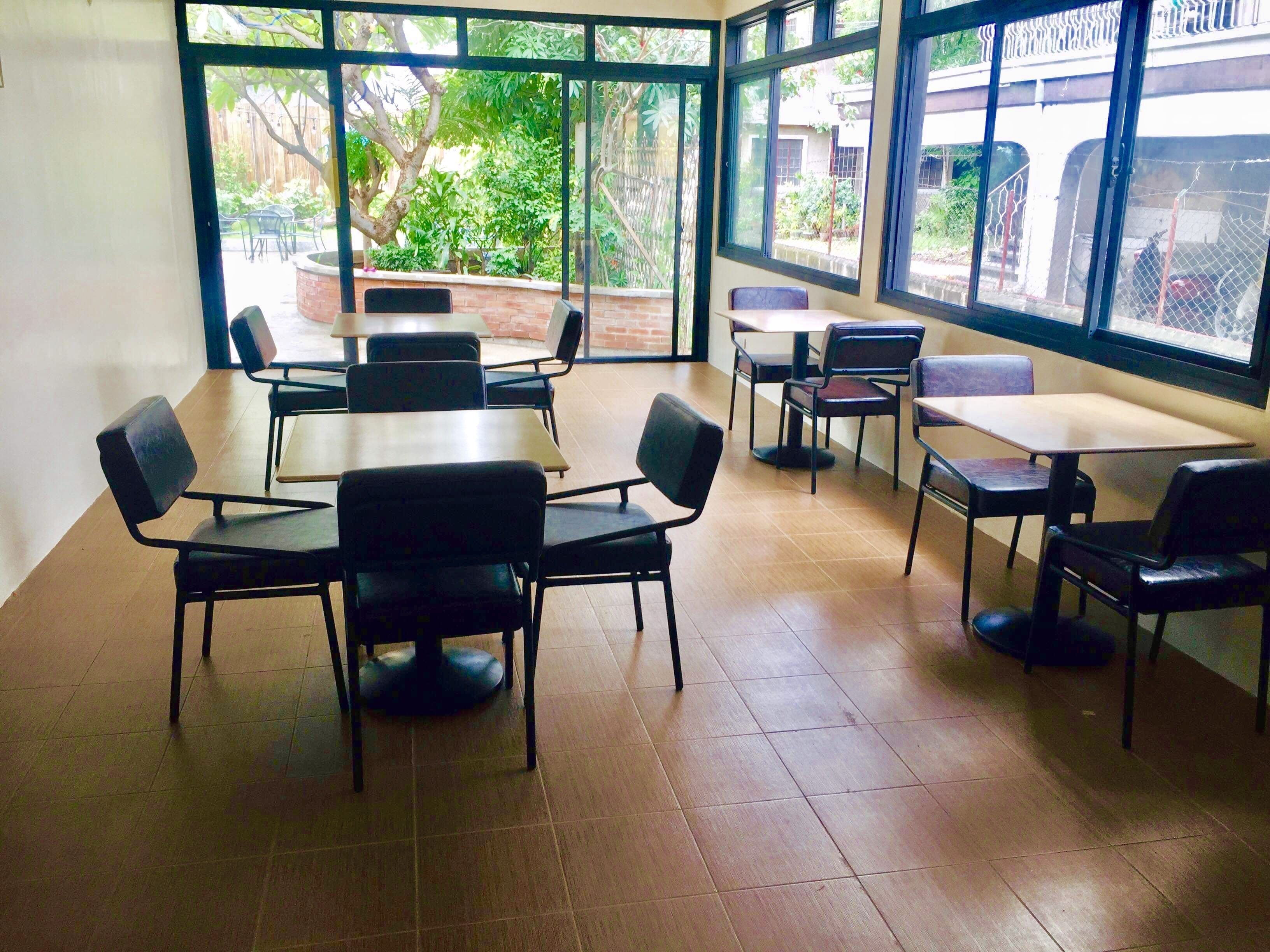 A spacious indoor area with multiple tables and chairs, large windows offering a view of a garden and buildings outside.