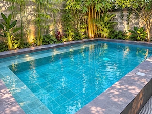 A rectangular swimming pool with clear blue water, surrounded by lush greenery and a stone wall. - The Hub District Garden Suites (Pasay)