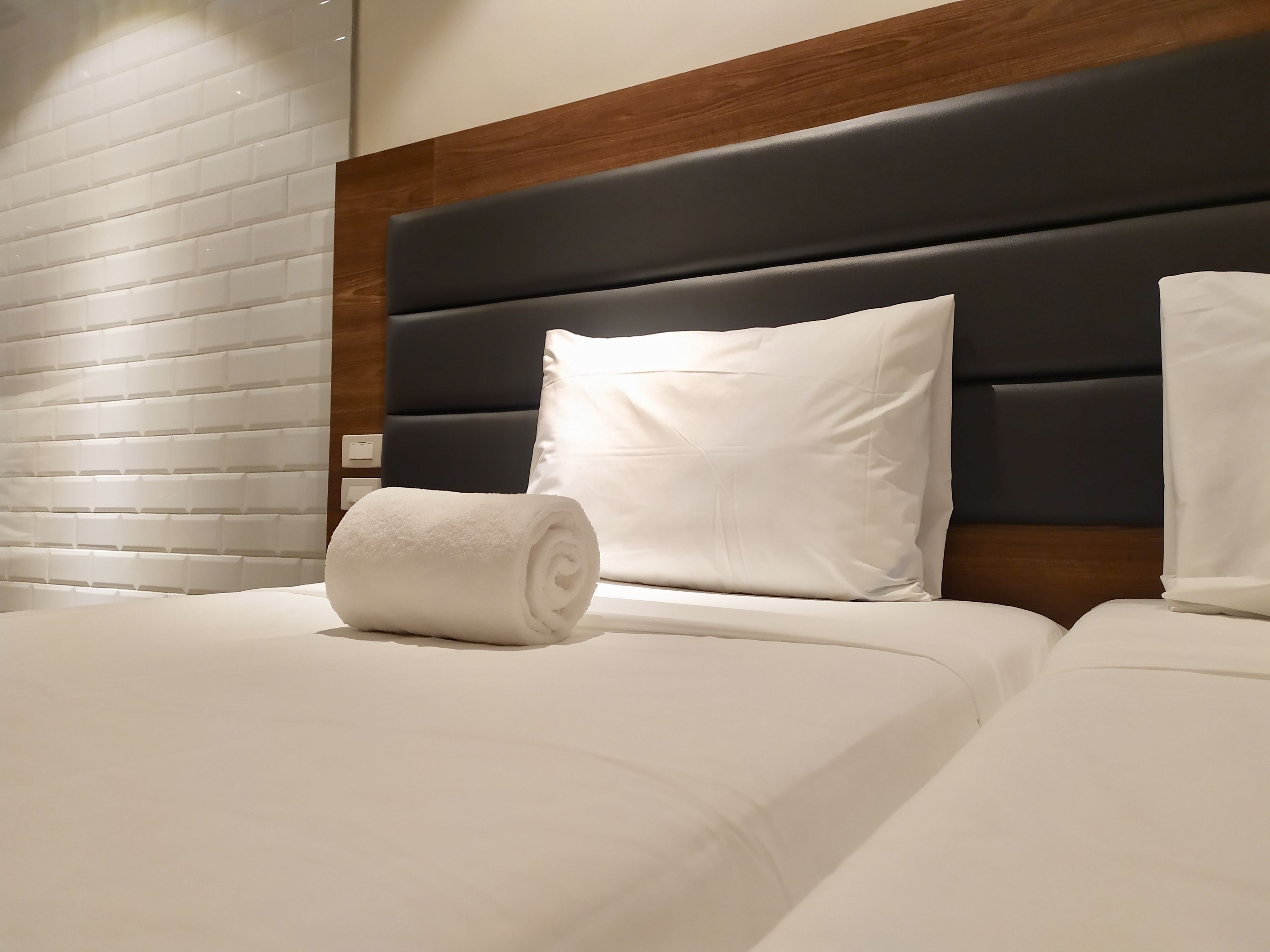 A neatly made bed with white linens and a rolled towel, a wooden headboard, and a white pillow.
