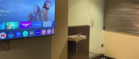 Quarto de hotel com televisão exibindo a interface de um serviço de streaming, uma cama, banheiro com pia e espelho.
