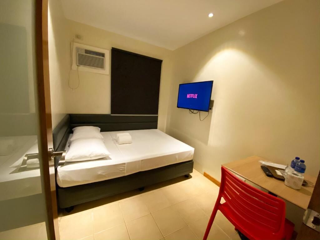 A hotel room with a bed, a desk with a chair, a TV mounted on the wall, and a window with blinds.