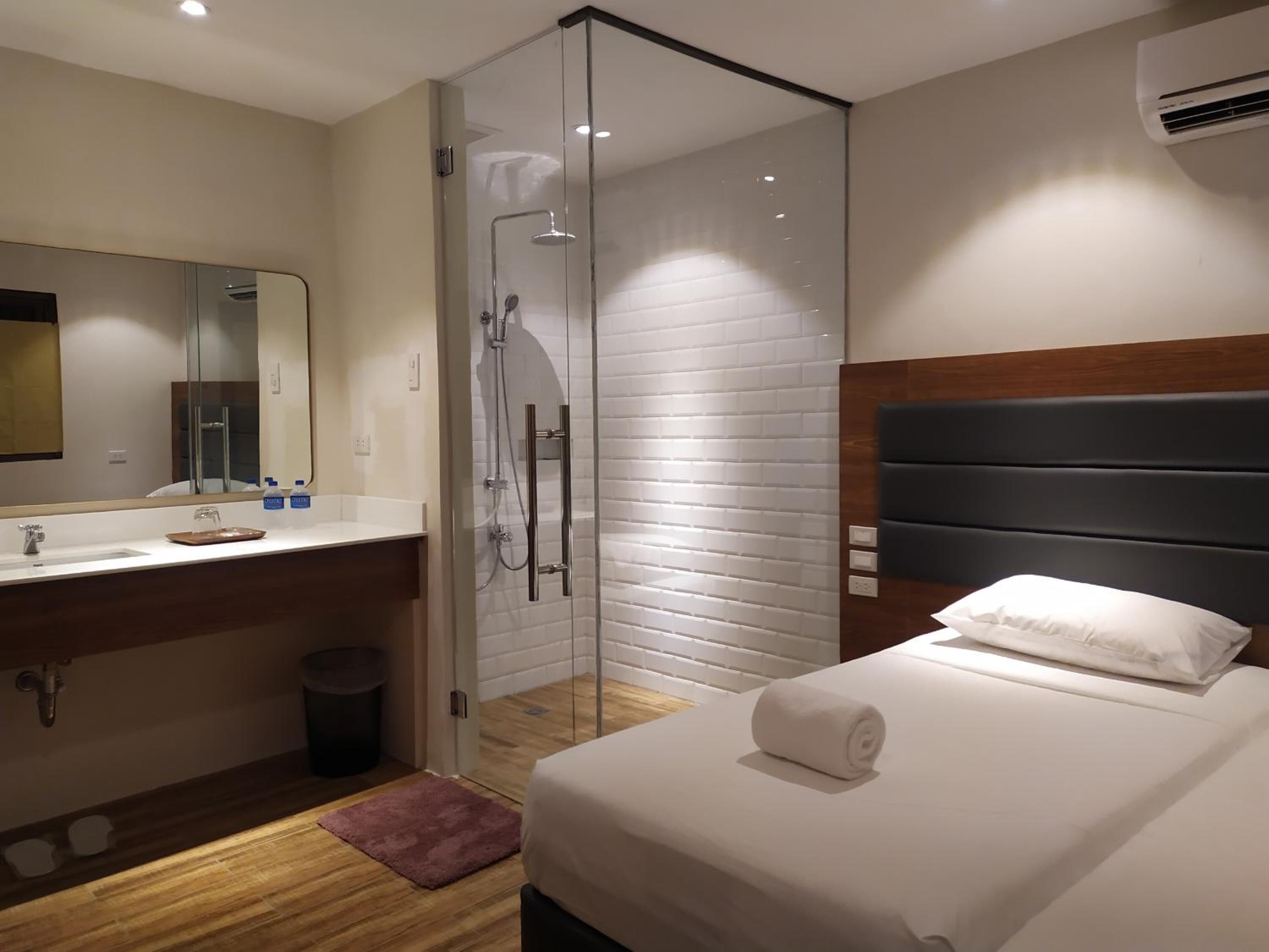 A modern hotel room with a glass-enclosed shower, a wooden floor, a large bed with a white pillow, and a wooden headboard.