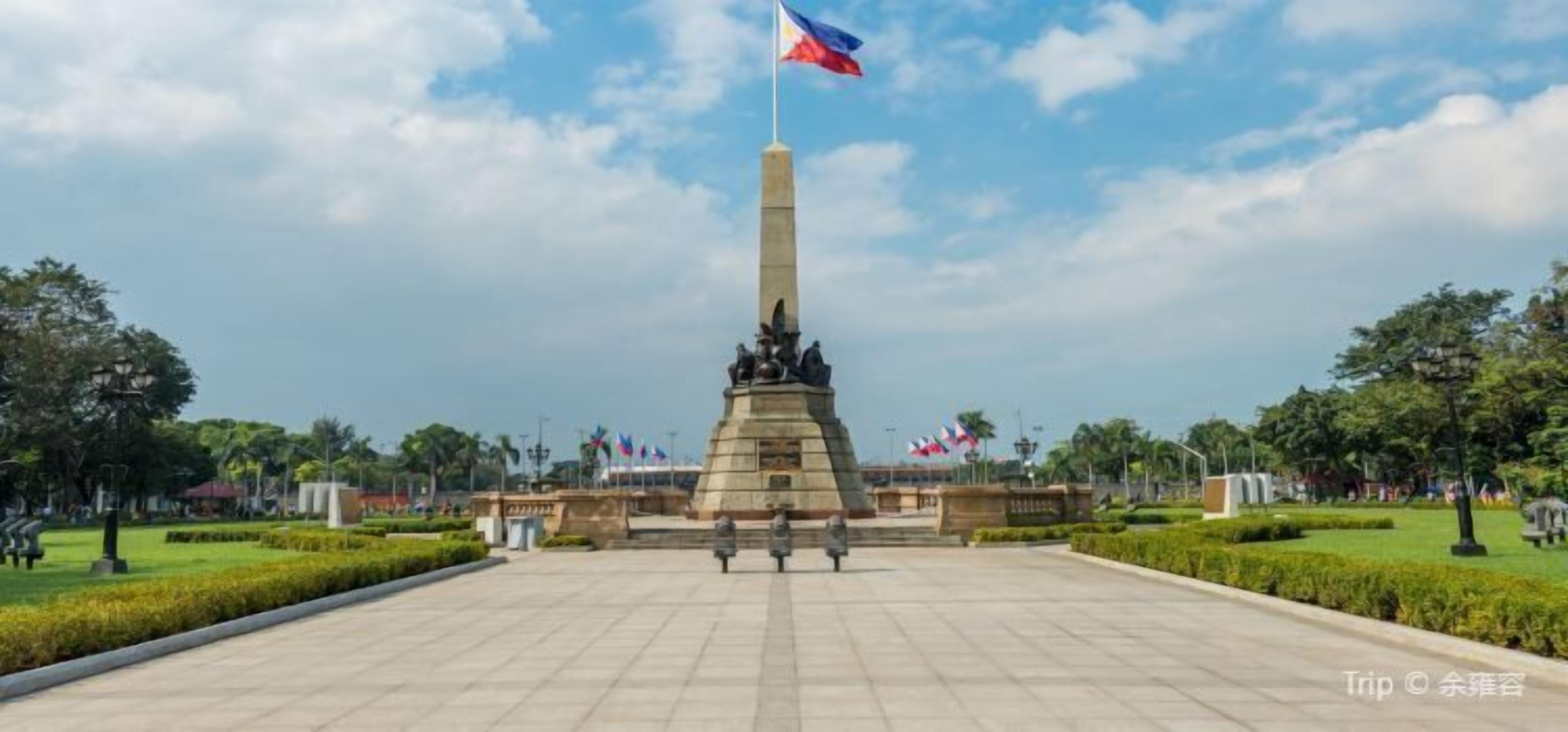 A monument with a flag, surrounded by a landscaped park with trees and a clear sky.
