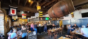 Un restaurante animado con zona de bar, vigas de madera y un barril colgante.