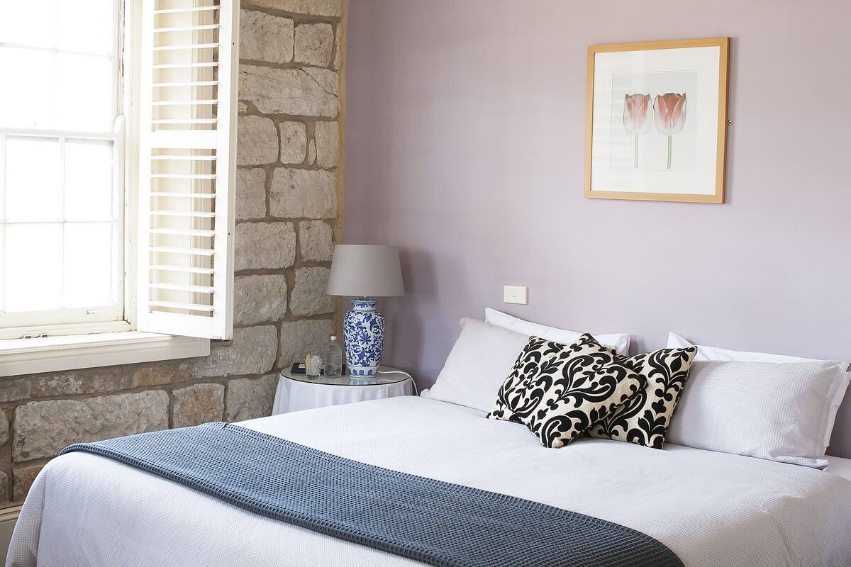 Un dormitorio con pared de piedra, una cama con almohadas estampadas, una mesita de noche con lámpara y una ventana con contraventanas.
