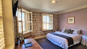 A bedroom with a large bed, two windows with shutters, a stone wall, a bedside table with a lamp, and a framed picture on the wall. - The Lord Nelson Brewery Hotel (Millers Point)