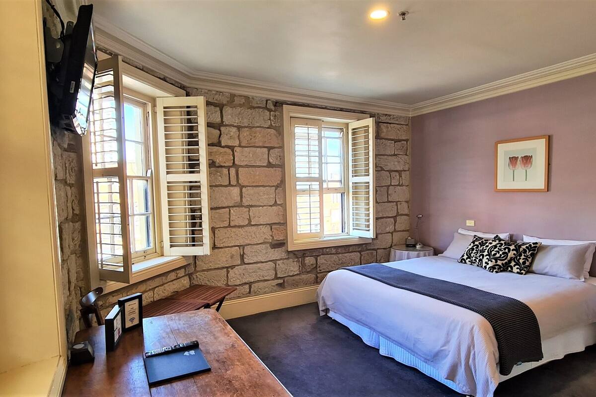Un dormitorio con cama, un escritorio, dos ventanas con contraventanas, un muro de piedra y un cuadro en la pared.