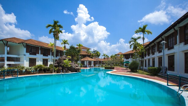 A large swimming pool surrounded by multi-story buildings with balconies, palm trees, and a clear blue sky. - Villea Morib (Banting)