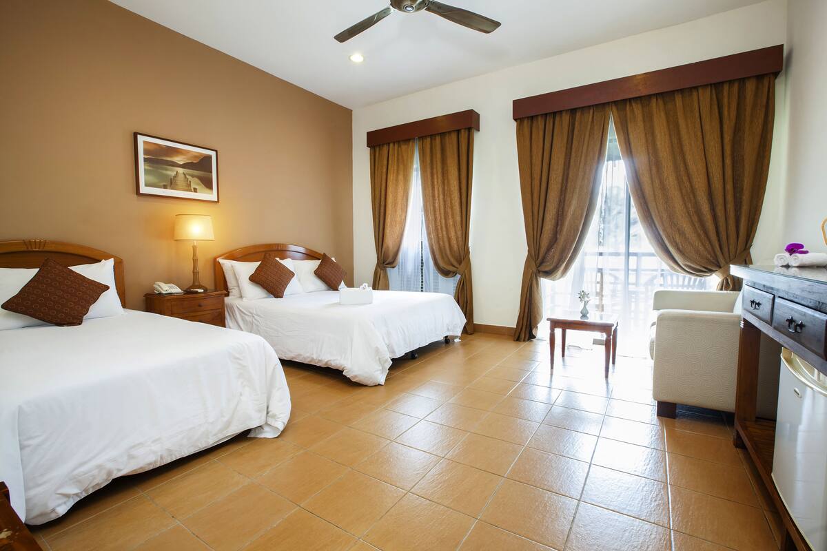A hotel room with two beds, a desk, a chair, a ceiling fan, and a window with curtains.