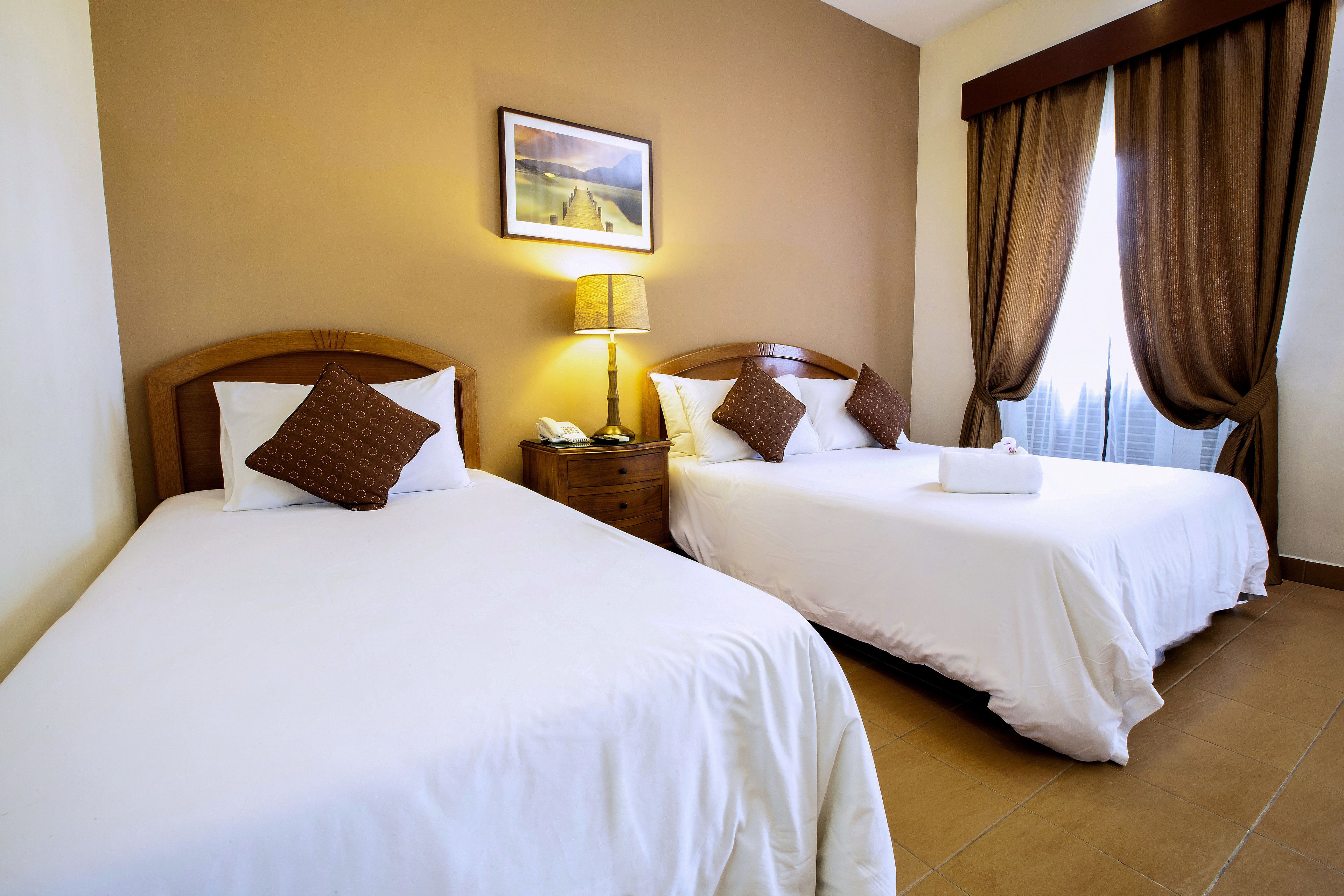 A hotel room with two beds, a nightstand with a lamp, a painting on the wall, and a window with curtains.