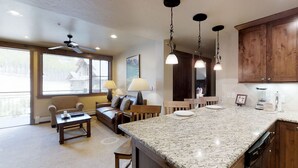 A spacious kitchen with granite countertops, wooden cabinets, and pendant lighting. There is a dining area with a sofa and a table, and a large window providing natural light. - 2 Bd Grand Lodge Peak 7 - sleeps 8 (Breckenridge)
