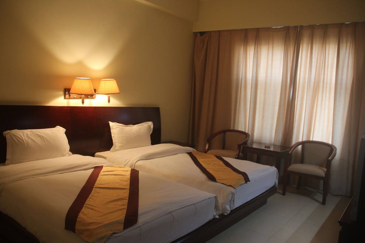 A hotel room with two beds, a desk, a chair, and two wall-mounted lamps.