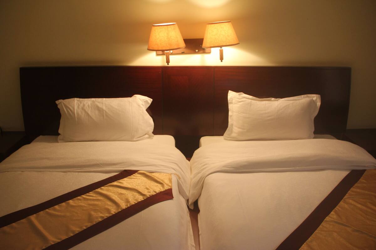 A hotel room with two single beds, a wooden headboard, and two wall-mounted lamps.
