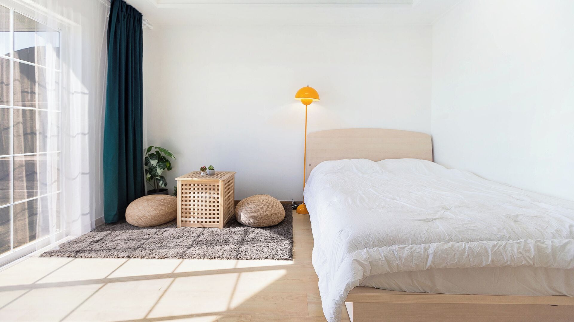 A bedroom with a bed, a floor lamp, a small table with a plant, and a window with curtains.