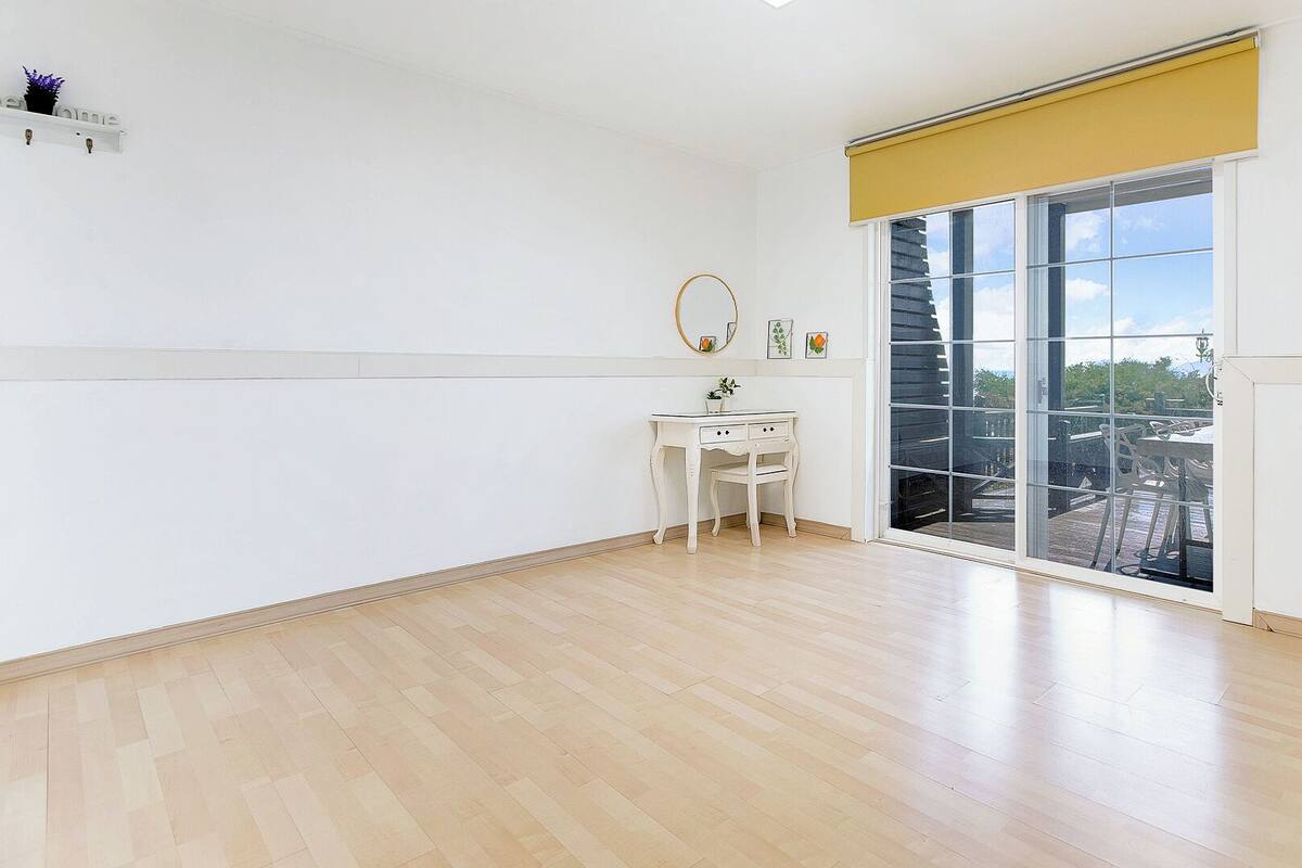 A spacious room with a large sliding glass door leading to an outdoor area, a small white desk with a chair, and a wall-mounted shelf with decorative items.