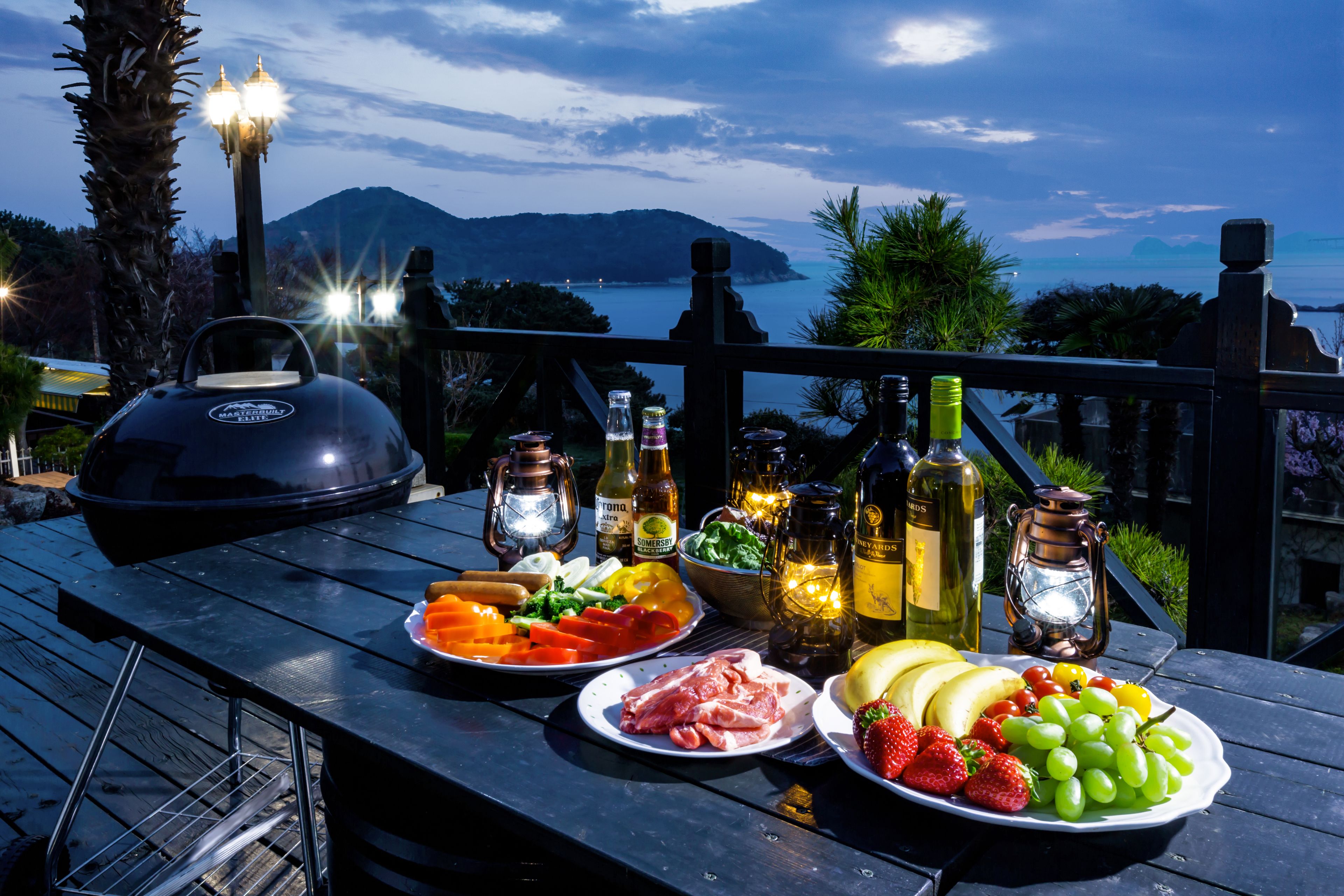 Una terrazza in legno con una griglia per barbecue, piatti di cibo e bottiglie di bevande, con vista panoramica su un lago e montagne.