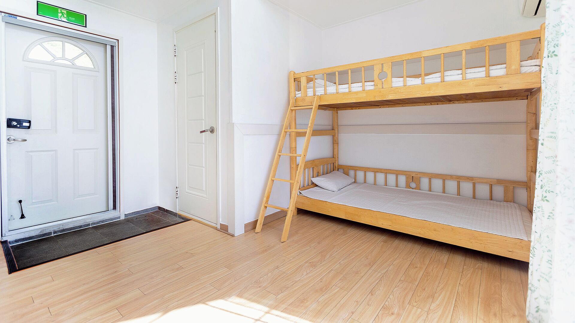 A bunk bed room with a wooden ladder, a white door with a green emergency exit sign, and a wooden floor.