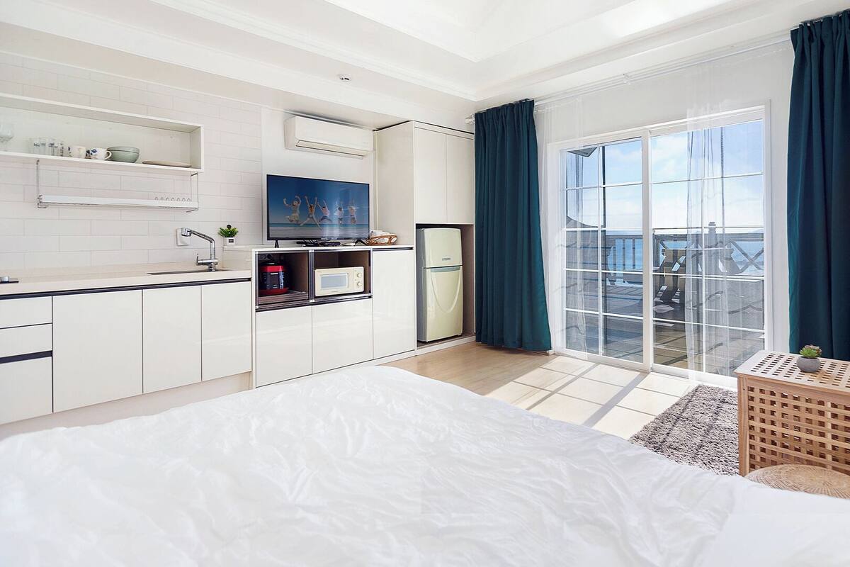 A modern hotel room with a large bed, a kitchenette, a balcony with curtains, and a view of the sea.