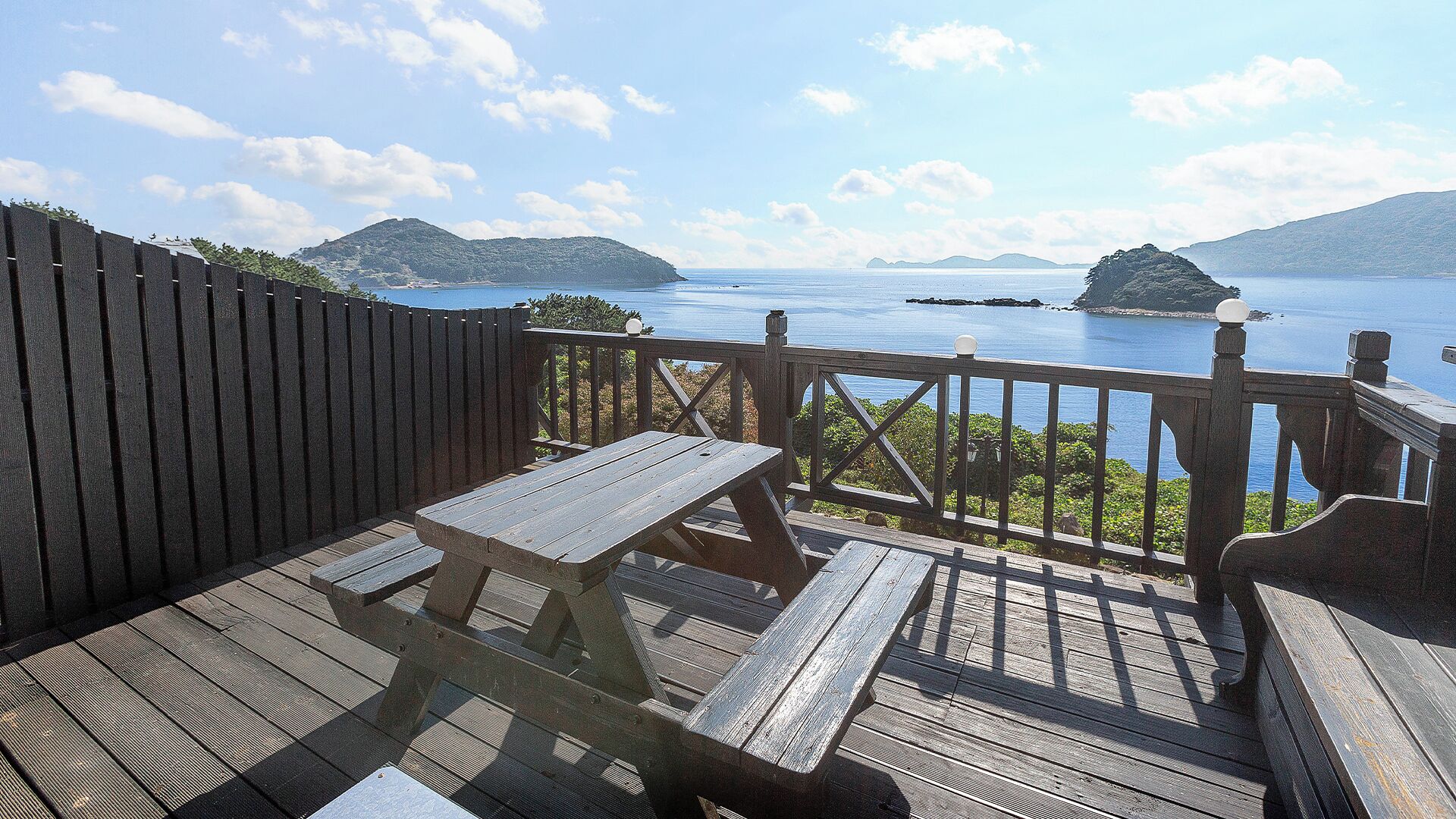 A wooden deck with a picnic table and benches, overlooking a calm body of water and distant islands.