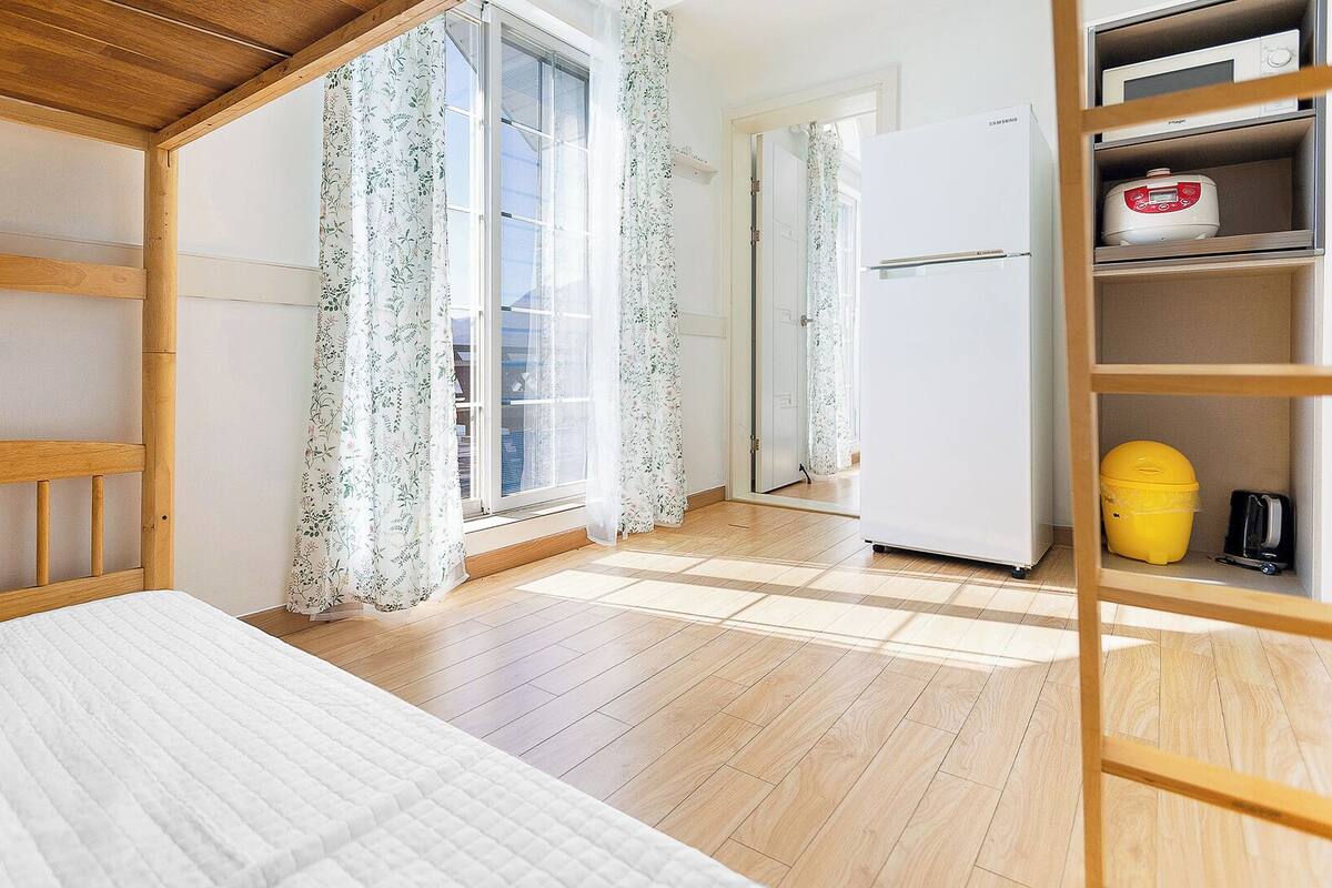 A room with a bed, wooden floor, a window with floral curtains, a white refrigerator, and a yellow bucket.