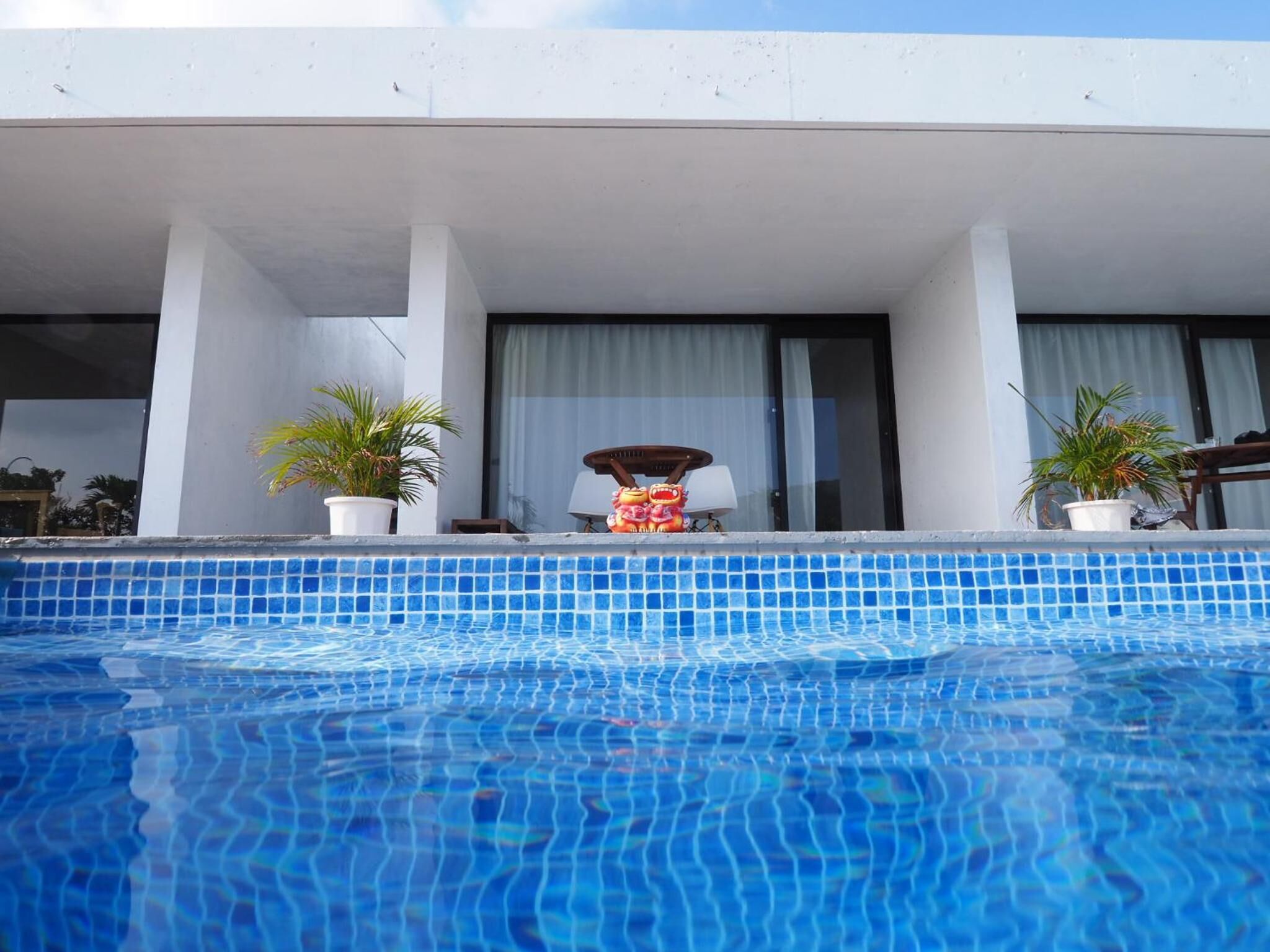 Una piscina con borde de azulejos, dos macetas, una silla y una mesa con lámpara.
