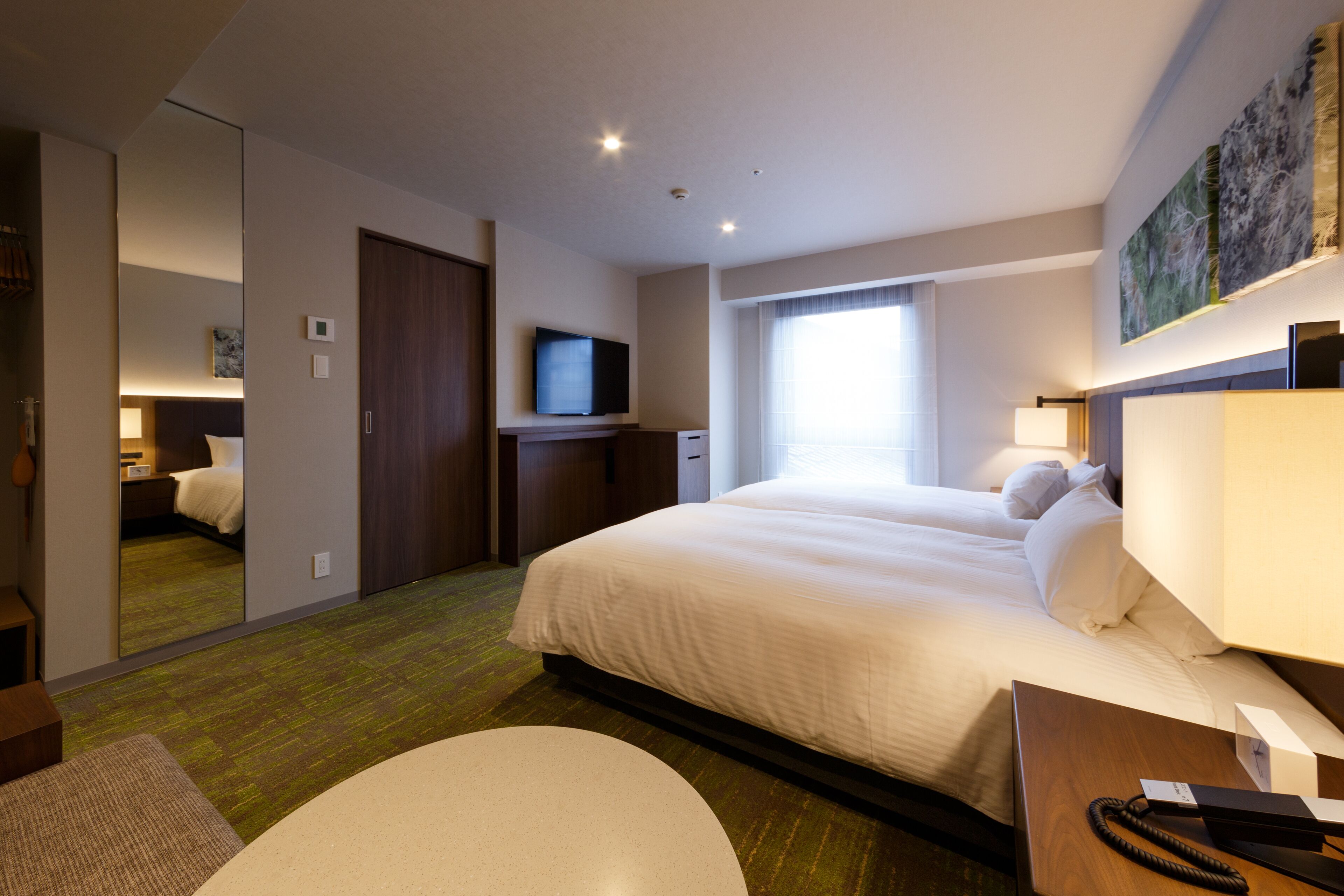 A hotel room with a large bed, a flat-screen TV, a desk with a lamp, and a window with curtains.