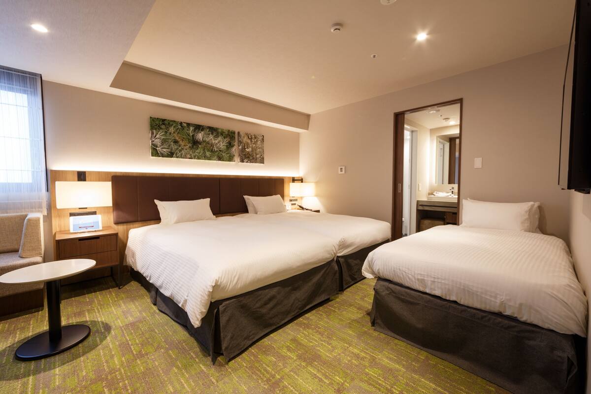A hotel room with two beds, a small table, a chair, and a large window.