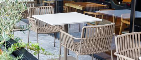 Outdoor seating with wicker chairs and white tables, surrounded by potted plants.
