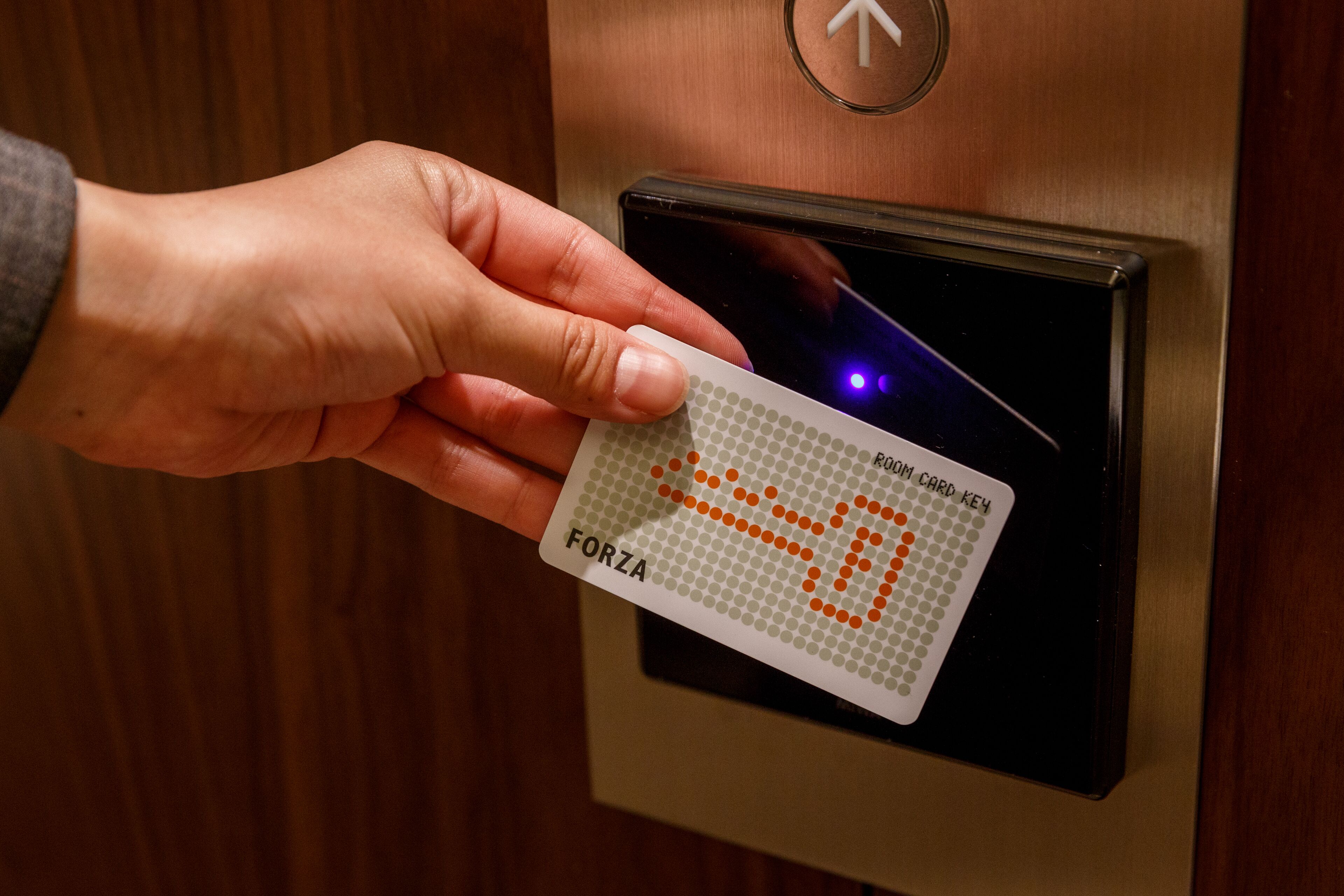 Une main tenant une carte ornée d’un motif à pois et portant le mot « FORZA », que l’on insère dans un lecteur de cartes situé sur la porte d’un ascenseur.