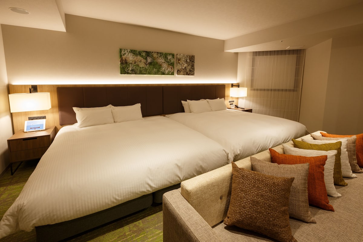 A hotel room with two large beds, a nightstand with a lamp, and a wall-mounted artwork.