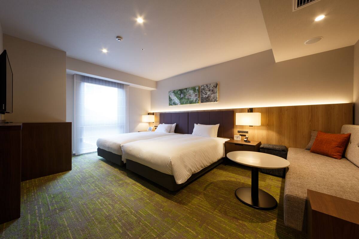A hotel room with two beds, a desk, a chair, a small table, and a TV.