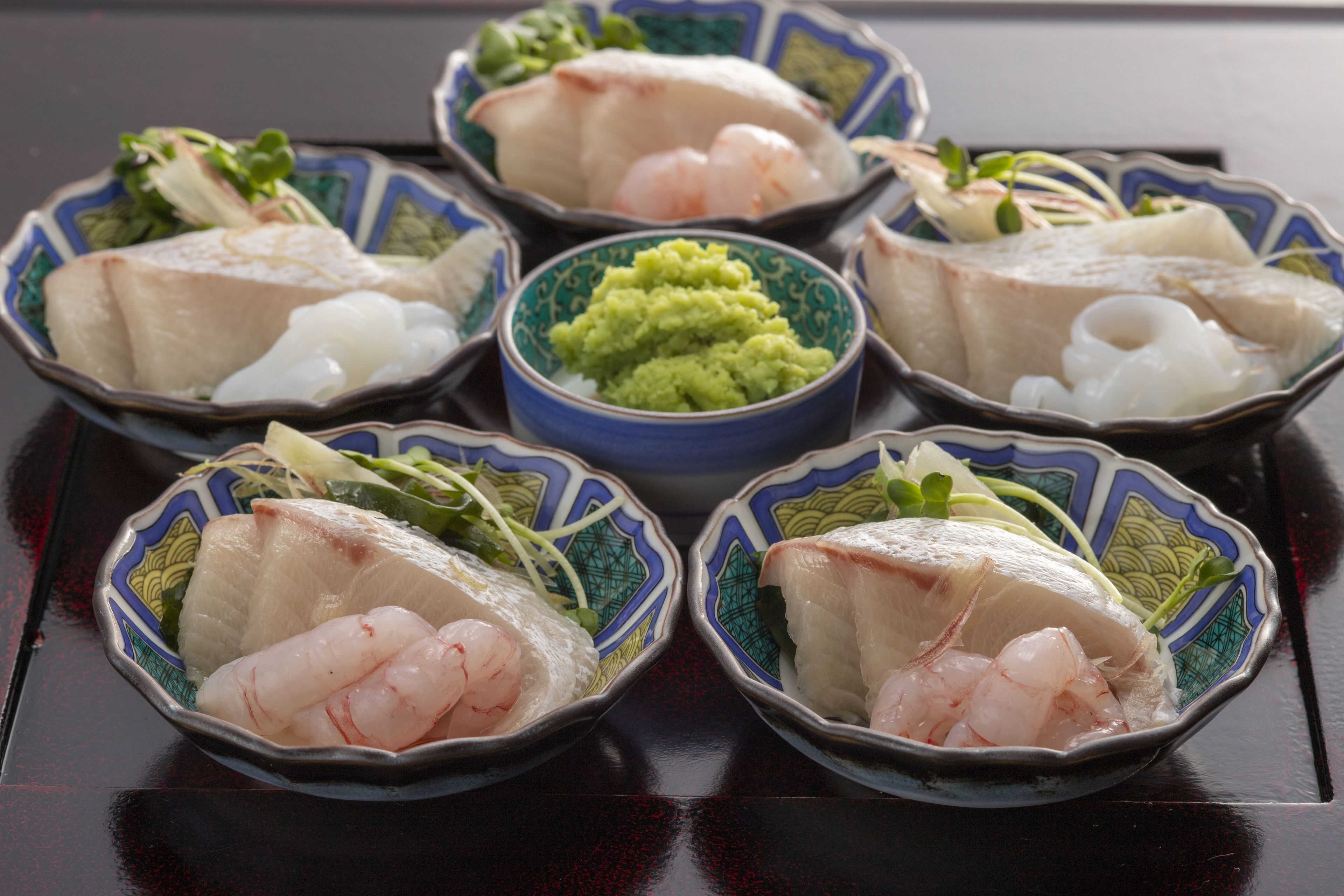 A table with six bowls of sashimi, each with different types of fish and garnishes, and a central bowl of green sauce.