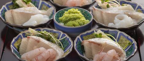 Una mesa con seis cazos de sashimi, cada uno con diferentes tipos de pescado y guarniciones, y un cazo central de salsa verde.