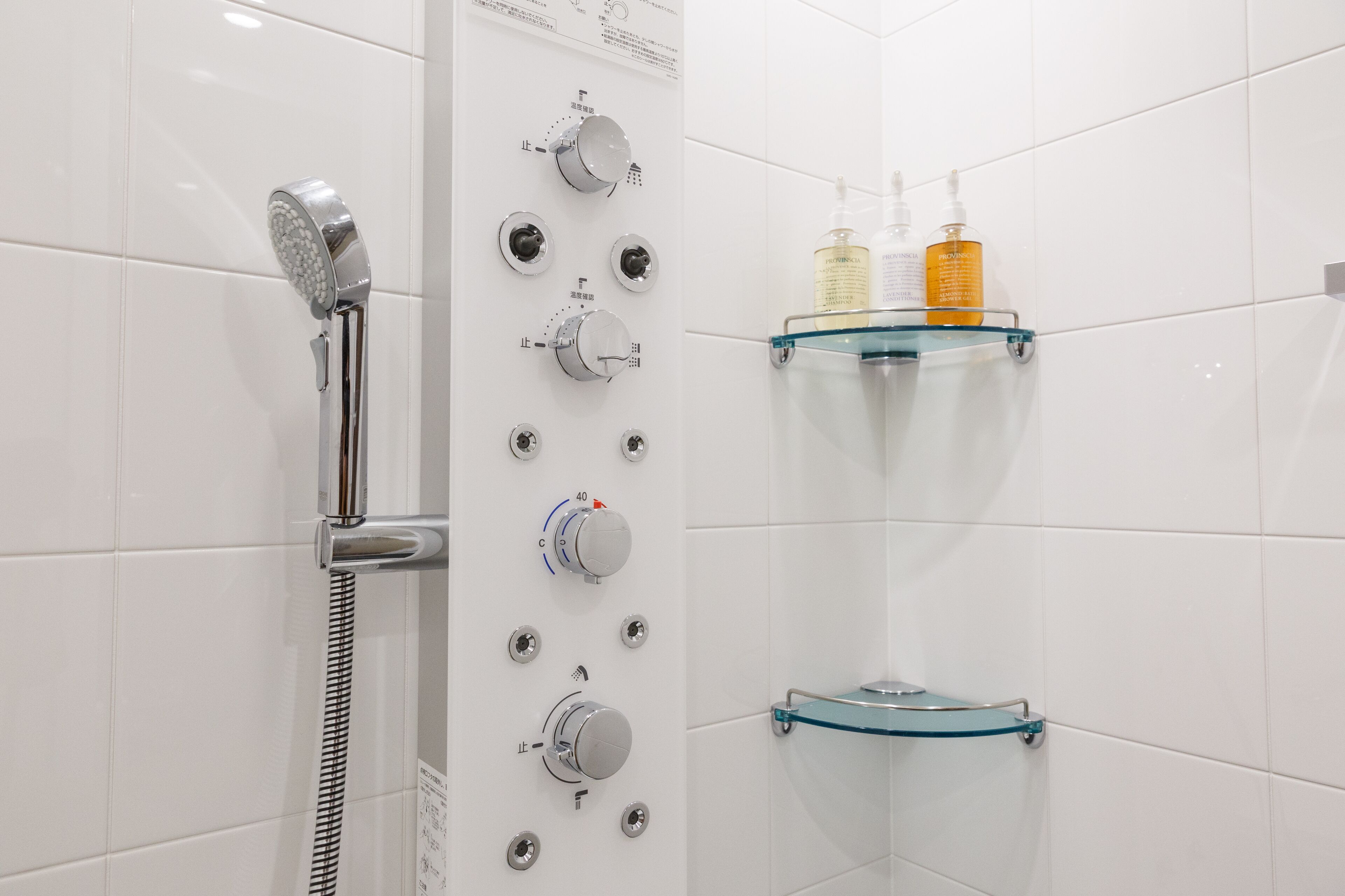 A modern bathroom with a glass shower enclosure, multiple control knobs, and a glass shelf with toiletries.