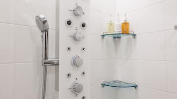 A modern bathroom with a glass shower enclosure, multiple control knobs, and a glass shelf with toiletries.