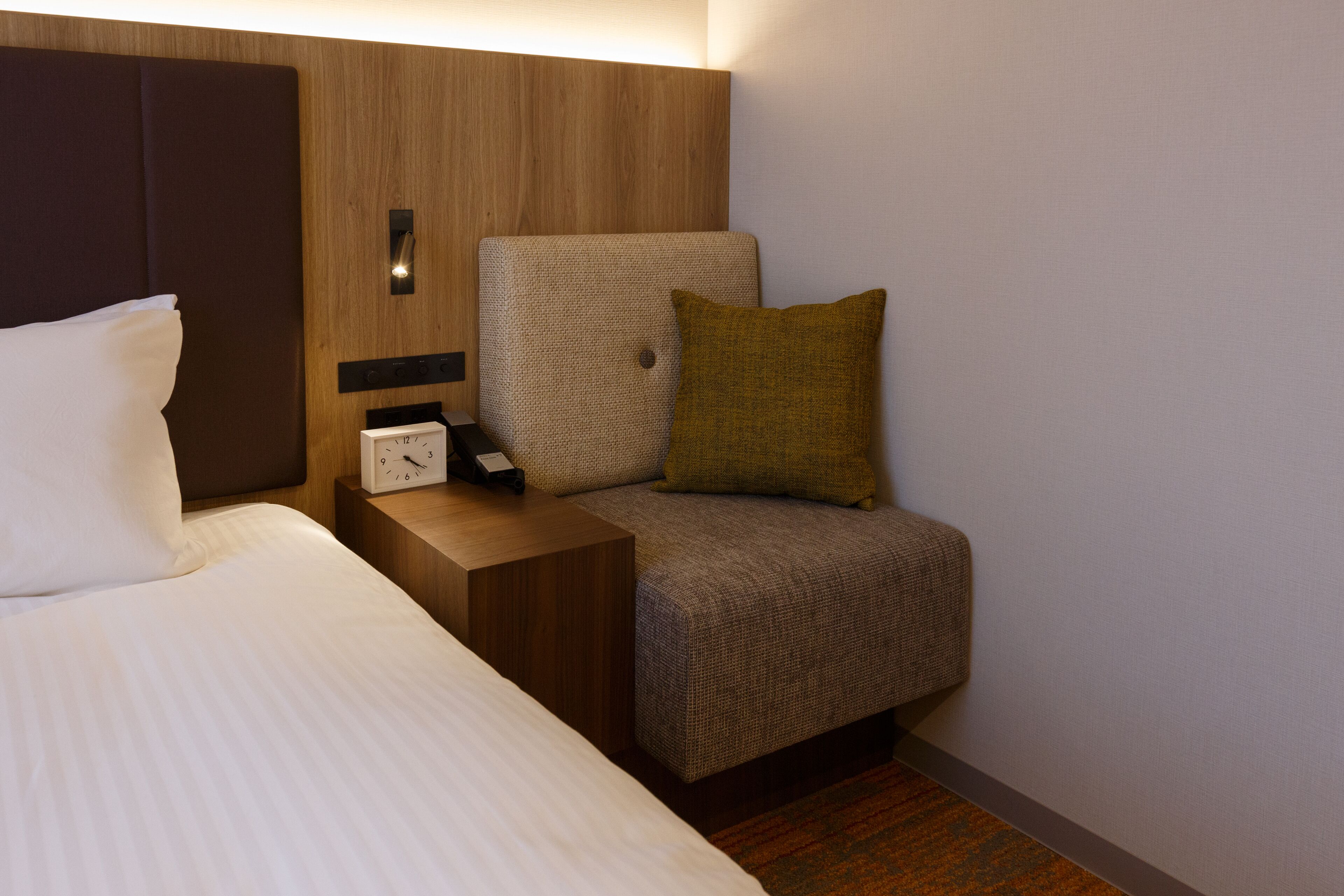 Ein Hotelzimmer mit einem Bett, einem Nachttisch mit einer Uhr, einem Sessel und einem Kopfkissen.