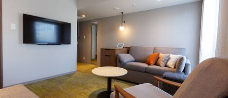 A modern living room with a flat-screen TV, a grey sofa with orange and white cushions, a wooden armchair, and a small round table.