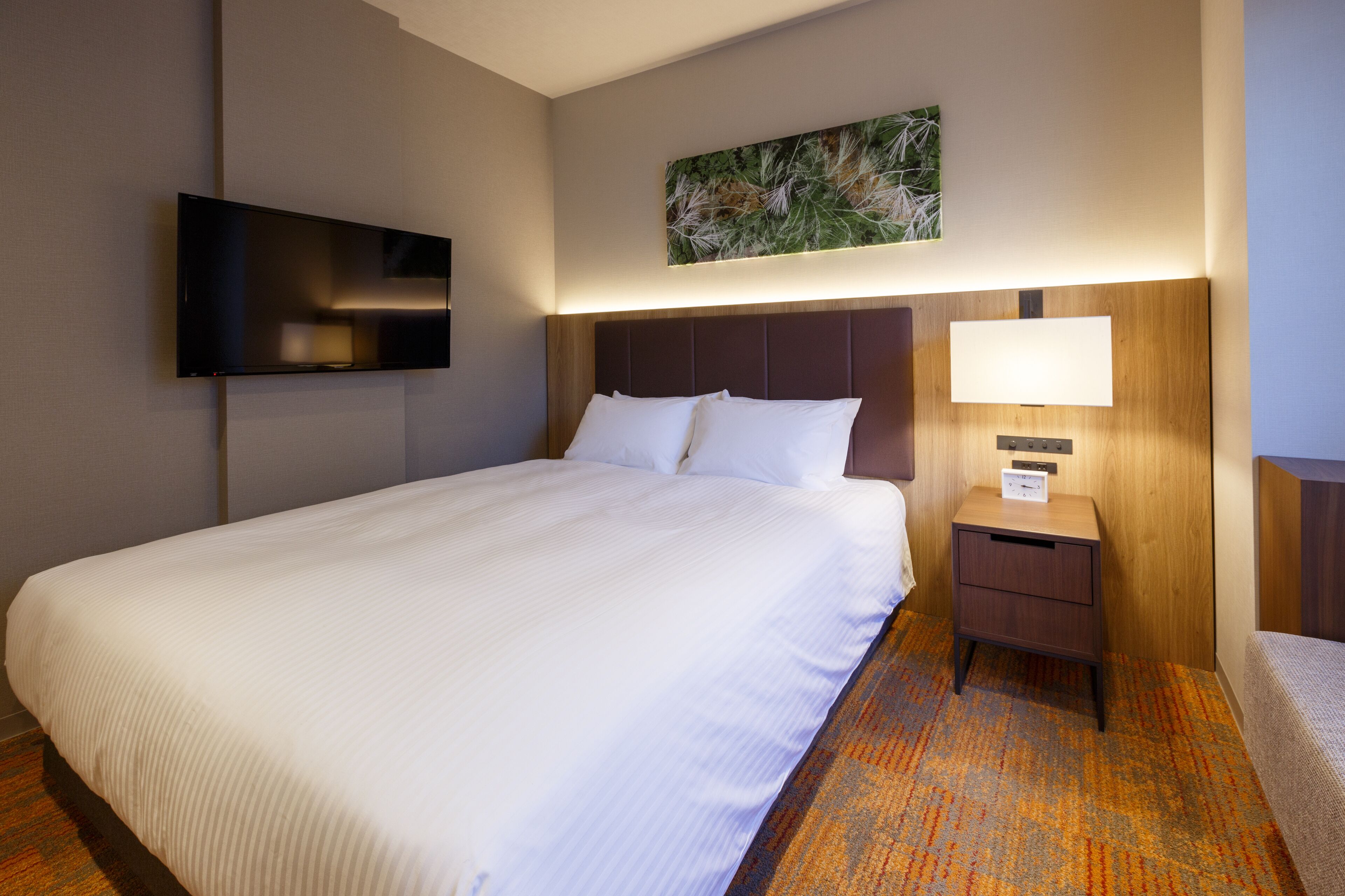 Une chambre d’hôtel avec un grand lit, un téléviseur à écran plat fixé au mur, une table de chevet avec une lampe et une petite œuvre d’art au-dessus du lit.